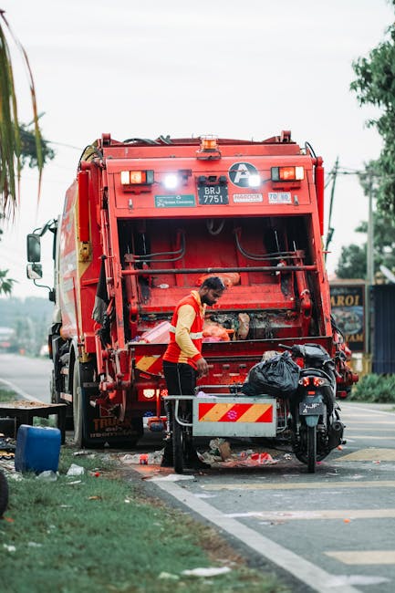 A waste collection worker dressed in a high-visibility yellow and red vest is operating a large, red rubbish truck on a roadside. The rear of the truck is open, revealing the internal compactor mechan