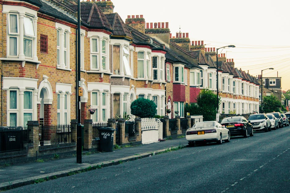 A row of traditional British terraced houses with brick facades and bay windows, many of which are painted in light pastel colors or left in natural brick tones, stand along a residential street scene