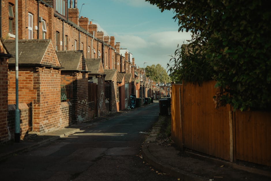 A row of traditional British terraced houses with brick facades and bay windows, many of which are painted in light pastel colors or left in natural brick tones, stand along a residential street scene